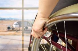 A close up photo of a person seated in a wheelchair, and their hand on the wheel is in close view, and in the background, through a window, is a parked airplane at an airport gate