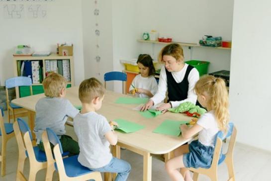 Teacher and young students sitting at a table folding and cutting green-colored construction papers