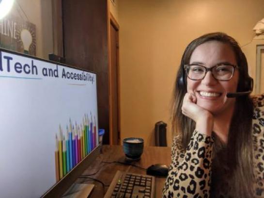 Dr. Fecich smiling by computer with a presentation on tech and accessibility